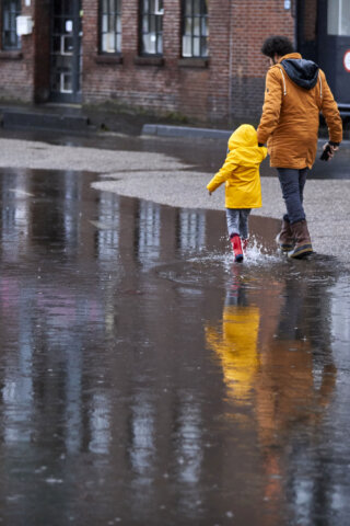 Een volwassene en een kind, gekleed in regenkleding, lopen door plassen water op een verharde straat.