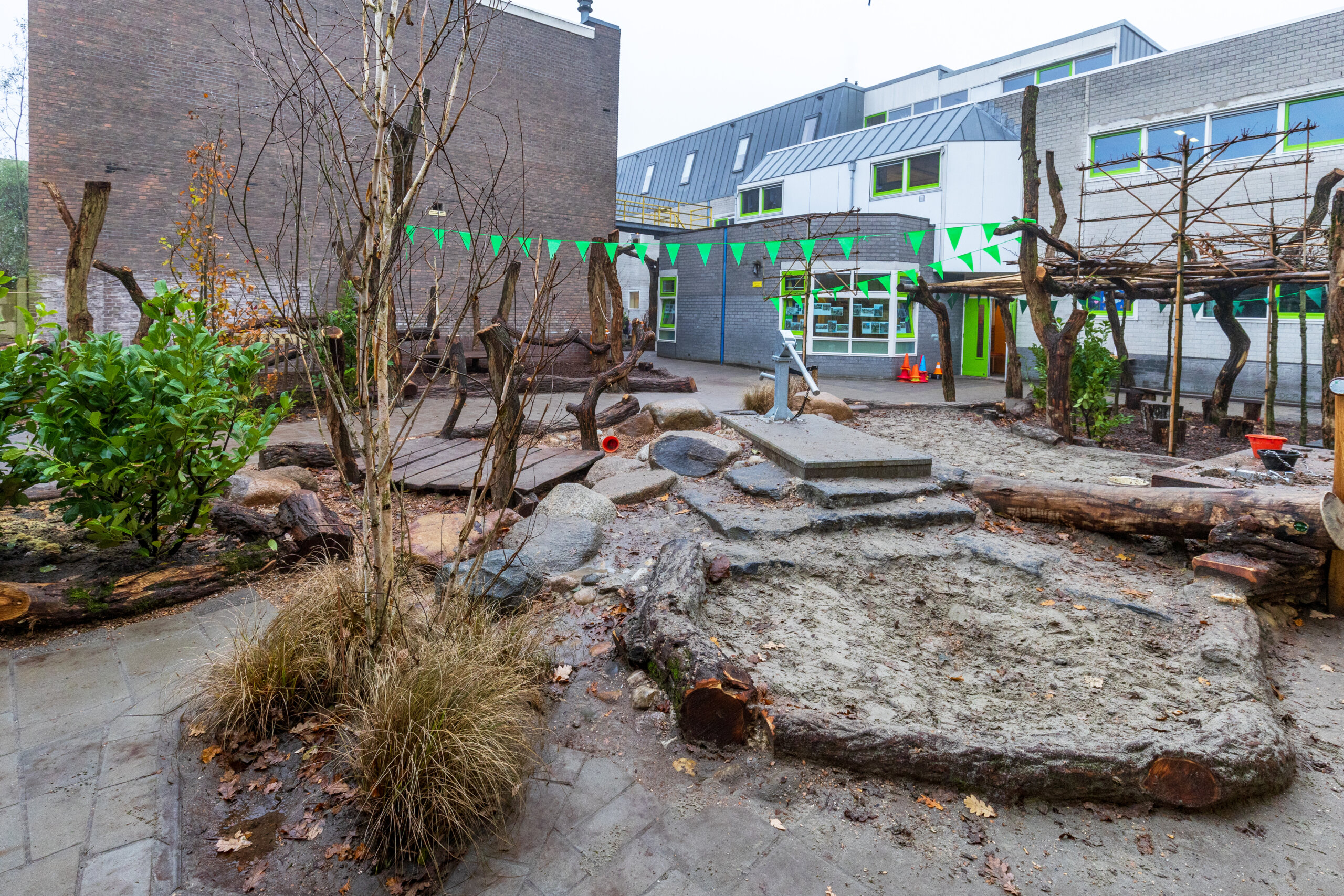 Een schoolplein met natuurlijke speelelementen zoals boomstammen. Er zijn planten en een waterpomp in het midden. Het is een open ruimte met gebouwen op de achtergrond.