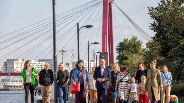 Een groep mensen van verschillende leeftijden loopt samen over een stoep met een rode brug op de achtergrond.
